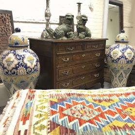 A vignette of antiques in an antique store contains a mahogany chest of drawers with brass pulls, two oversized covered ginger jars with Moroccan or Middle Eastern design motifs in blue, yellow, green, and white, a pair of green marble-like stone carved f | BRG