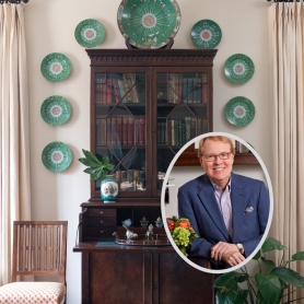 Interior designer, John Marrs is in an inset photo over a styled vignette consisting of an antique secretaire surrounded by by decorative green Transferware plates hanging on a wall. | BRG