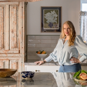 Emily stands in her kitchen next to a rustic cabinet and in front of a vintage still life painting. | BRG