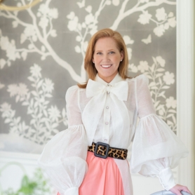 Portrait of smiling Lindsey standing in a dining room, wearing a white puff-sleeved shirt, light pink skirt, and a leopard-patterned belt. | BRG