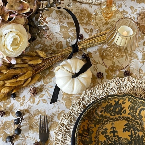 A gold and black dish on a woven place mat, a white pumpkin with black ribbon, dried floral bouquet, and amber glassware on a floral tablecloth help set the tone for this fall placesetting. | BRG