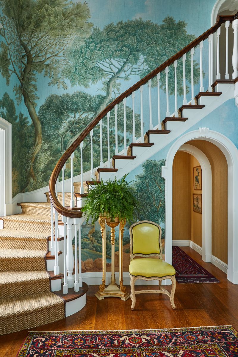 A luxurious foyer and sweeping curved stairwell adorned with mural wallpaper, featuring an antique carpet, a regal yellow chair, and an indoor plant on an elegant stand