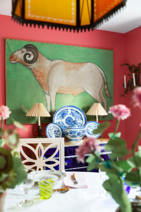 A boldly colored dining room with watermelon toned walls, a brightly painted ram painting, blue and white porcelains, blue chest of drawers, chairs and dining table set with dishes, colorful glassware and plants. 