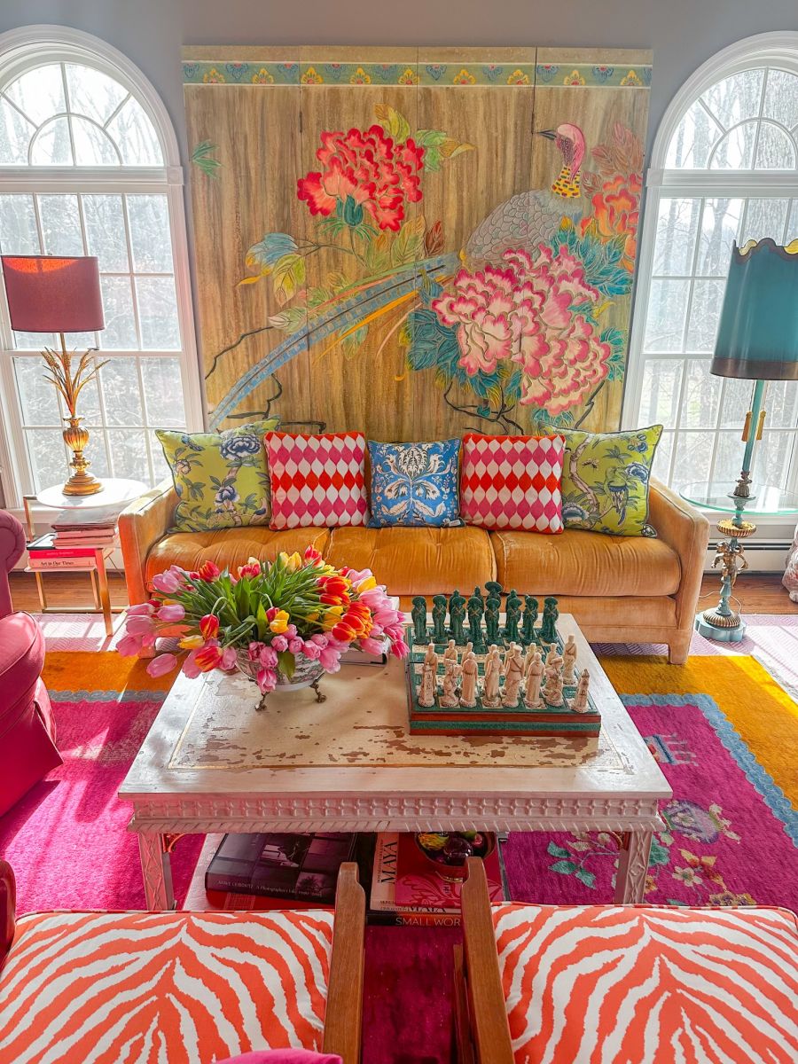 Color and pattern fill this bright room with a floral tapestry hanging on a wall flanked by two Palladian windows. A gold/amber velvet sofa covered with colorful pillows, a distressed white painted covffee table and two orange and white zebra-print upholstered chairs sit upon a hot-pink, turquoise, and gold area rug