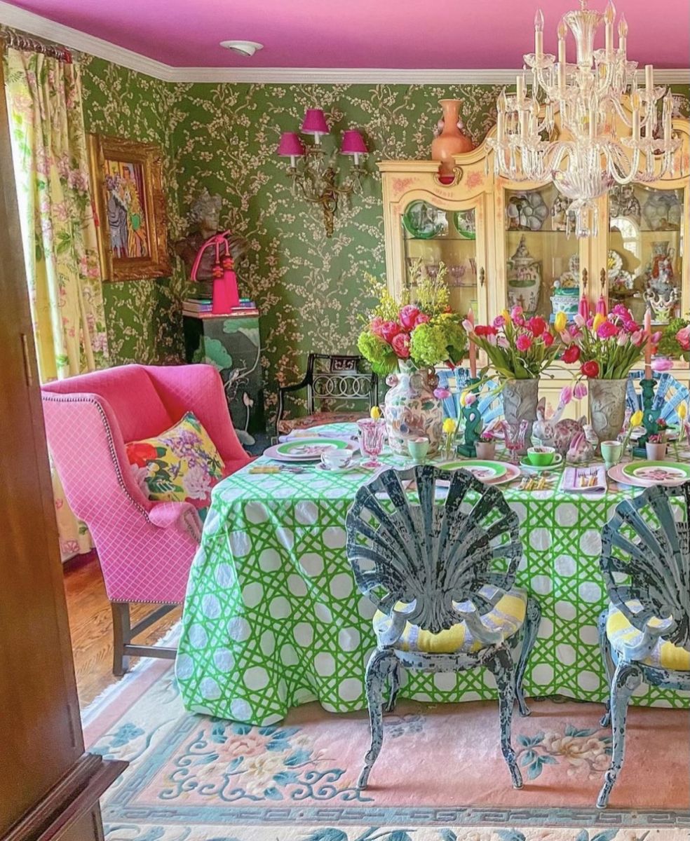 Vibrant colors and patterns rule this dining room from bold pink ceiling to green floral print wallpaper, opulent crystal chandelier, green and white geometric tablecloth and pastel blue, pink and cream Oriental rug.