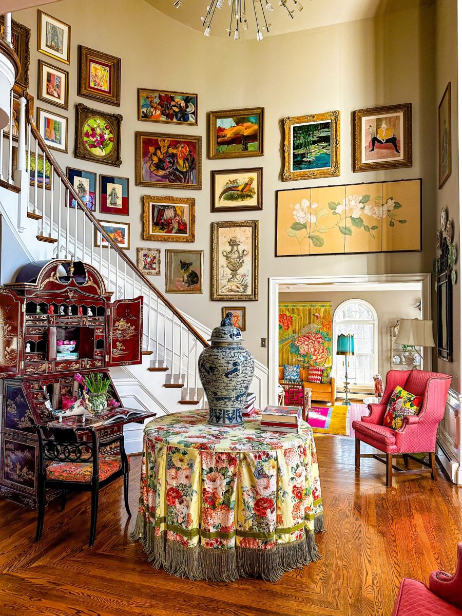 A grand foyer with a curved staircase and high ceilings features an extensive gallery wall filled with a variety of paints and works of art and a large round center hall table covered with floral patterned tablecloth and other Chinoisserie inspired pieces.