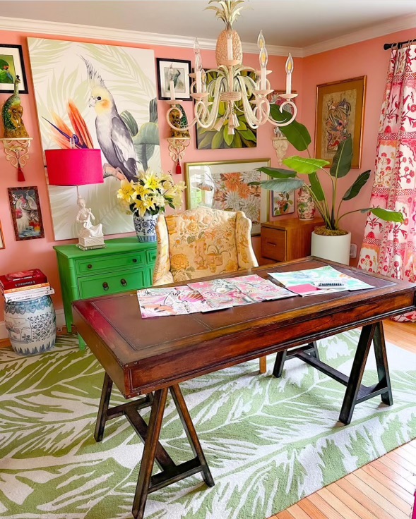 A vibrant workspace filled with intriguing details that spark inspiration during work hours, such as a painting of a parrot on the wall or a wingchair adorned with floral patterns in shades of yellow. This reflects Andrea's design style, characterized by a harmonious blend of bright colors, with pink and green as the predominant tones. Examples include a green dresser, lamp shades, window curtains, and walls painted in various shades of pink.