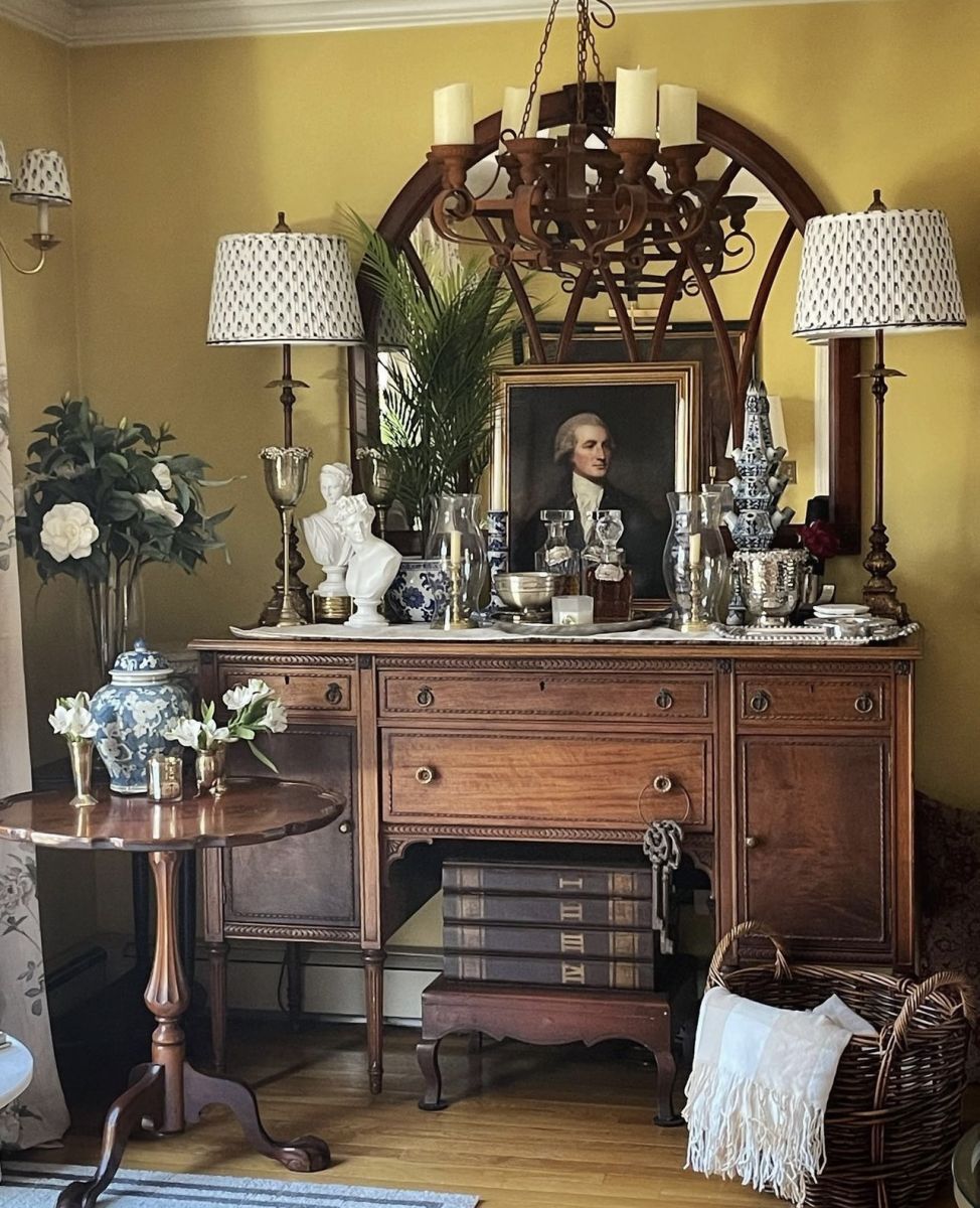 An antique server cabinet with intricate carvings and a weathered finish supports a pair of graceful table lamps with pretty blue and white patterned fabric shades. A large ornate mirror hangs on the wall above the server. An array of decorative accessories including delicate porcelain busts, sterling silver bowls and urns, and assorted blue and white porcelains add decorative style to the space