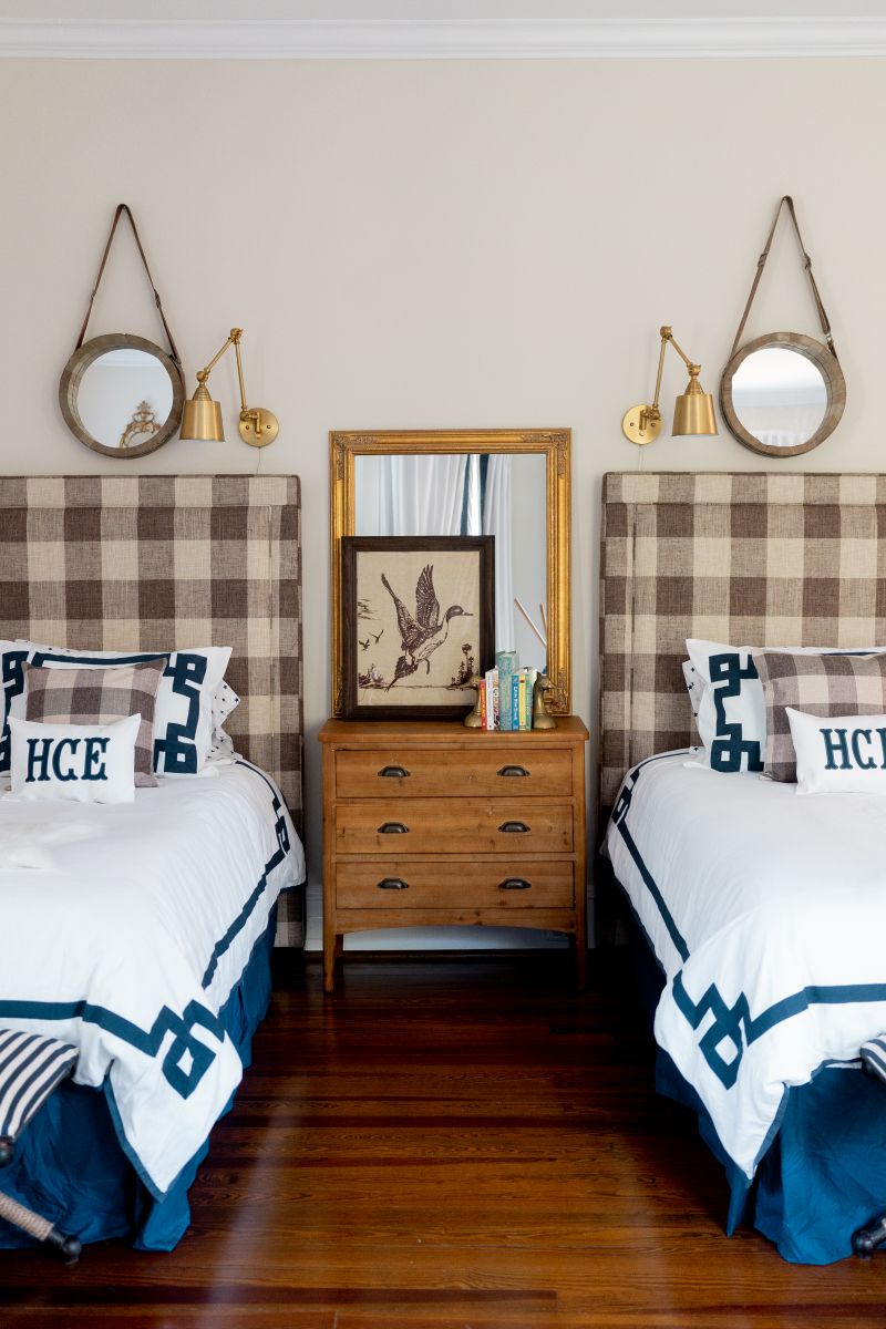 A twin bedroom mix and matches vintage items such as a brown wood chest of drawers / dresser used as a night stand, brass bookends and sconces, wooden framed mirrors with modern details such as pillows and blankets.