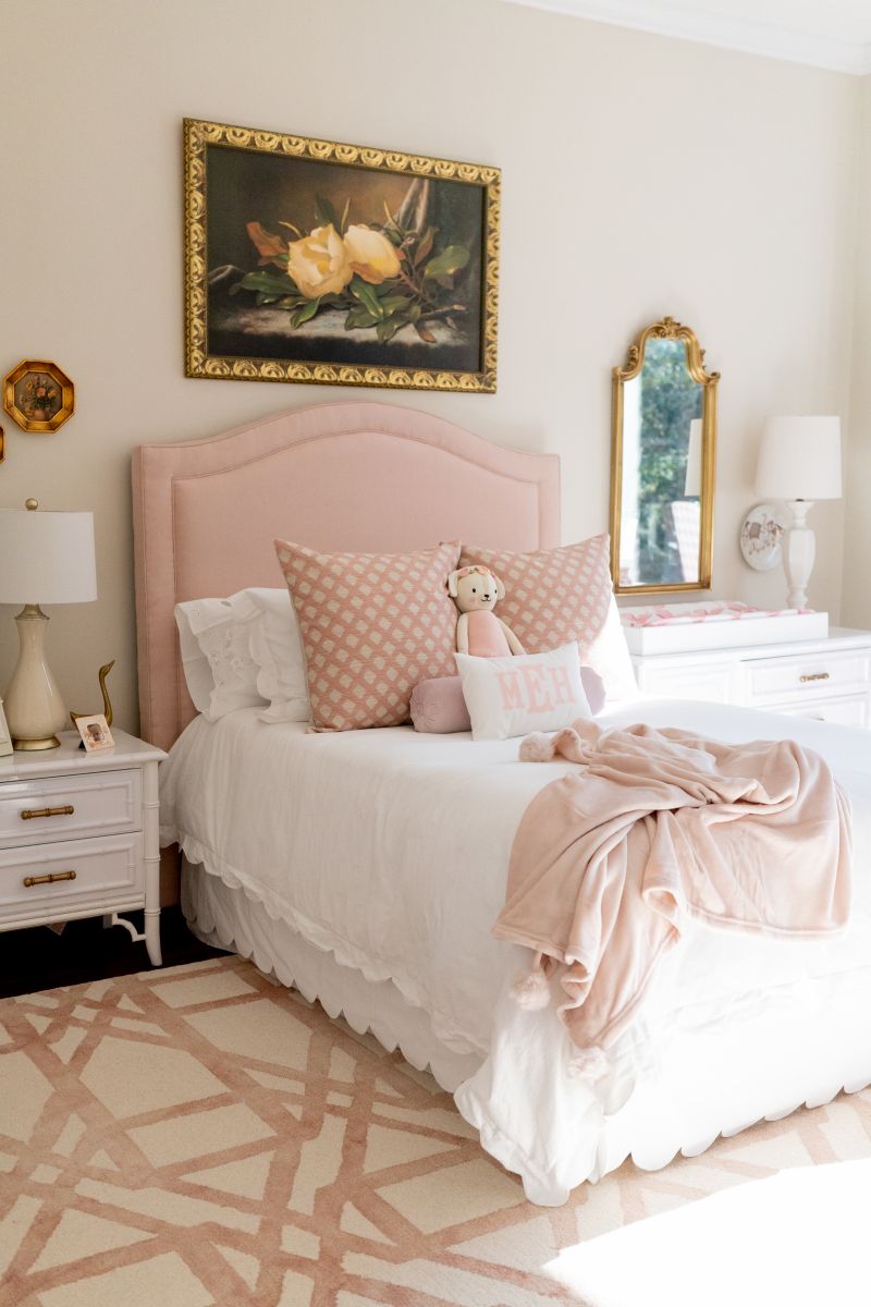 A pretty bedroom features vintage and timeless pieces in soft whites, creams, and pinks stand the test of time. Decoration includes a still life floral painting above the bed, gilt mirror and decorative accessories.