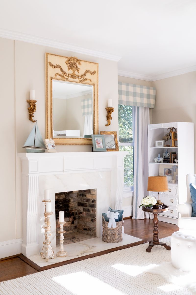 A designed fireplace mantle vignette features a huge gilt mirror, candlesticks and basket on the hearth, and several antique brown and soft muted white furniture pieces.