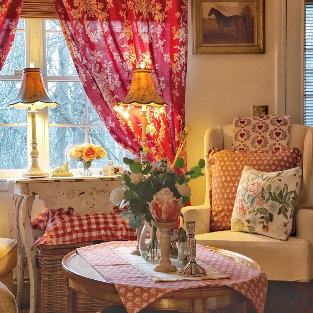 A comfortable upholstered armchair is adorned with a plush cushion and a knit throw blanket while natural light filters through a curtain a room with a pair of vintage-inspired table lamps creating a warm country French vibe.