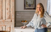 Emily stands in her kitchen next to a rustic cabinet and in front of a vintage still life painting.  | BRG